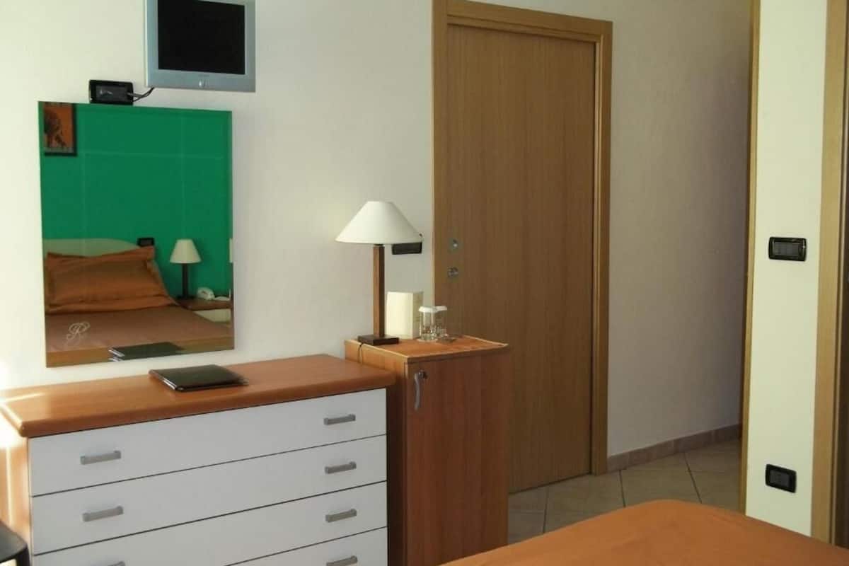 Un dormitorio con cama, cómoda con cajones, mesita de noche con lámpara, espejo, televisor montado en la pared y una puerta de madera.