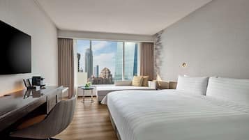 A modern hotel room with a large bed, a desk with a chair, a TV, and a view of the cityscape through large windows.