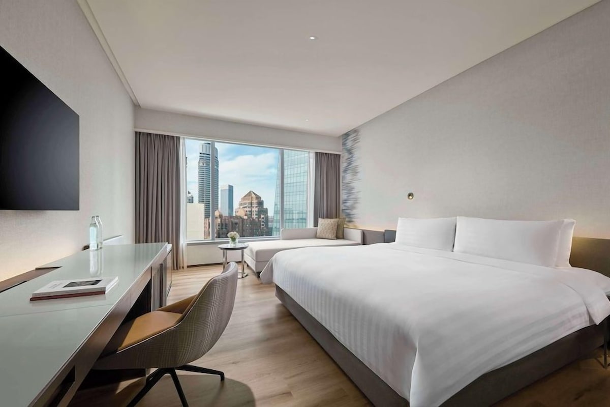 Ein modernes Hotelzimmer mit einem großen Bett, einem Schreibtisch und einem Stuhl. Ein Fenster bietet Blick auf die Stadt.