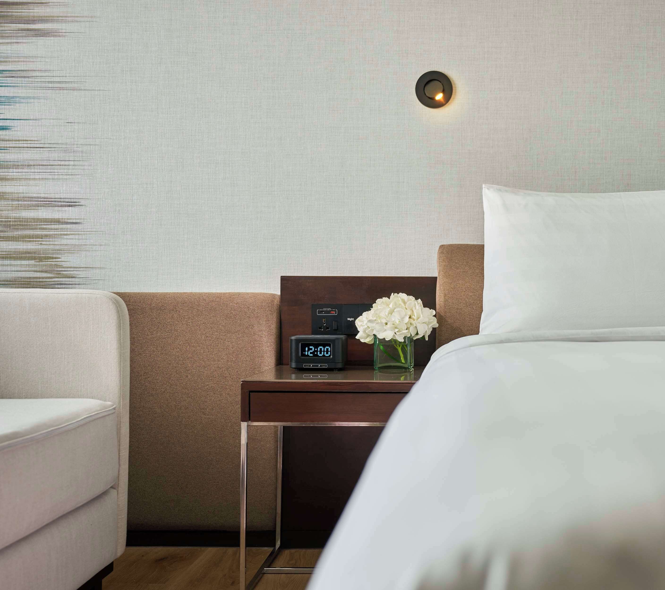 Una habitación de hotel con una cama, una mesita de noche con reloj digital, un florero con flores blancas y una lámpara fijada a la pared.