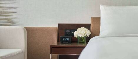 Una habitación de hotel con una cama, una mesita de noche con reloj digital, un florero con flores blancas y una lámpara fijada a la pared.