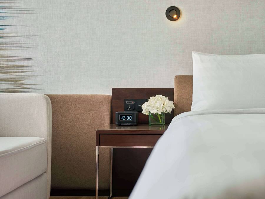 A hotel room with a bed, a nightstand with a digital clock, a vase with white flowers, and a wall-mounted light fixture.