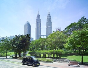 A park with two prominent skyscrapers in the background, a paved walkway, and a small vehicle.
