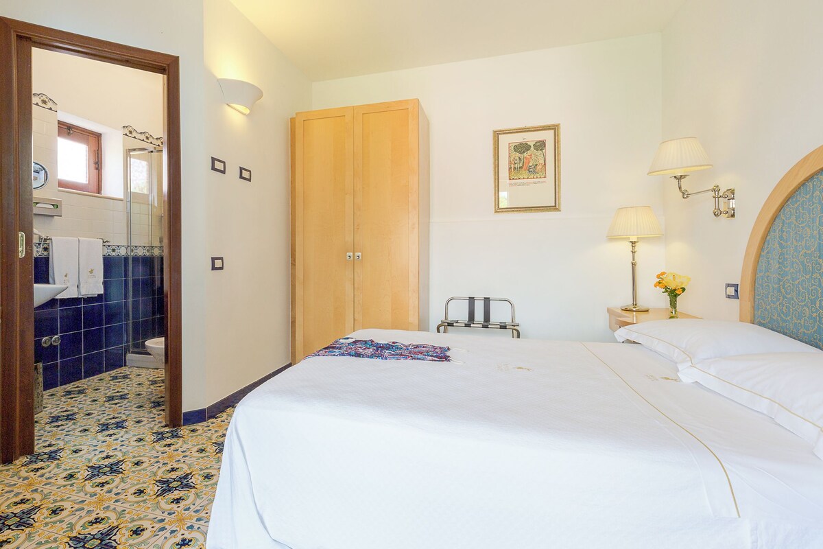 A hotel room with a bed, a wooden wardrobe, a nightstand with a lamp, and a bathroom visible through an open door.