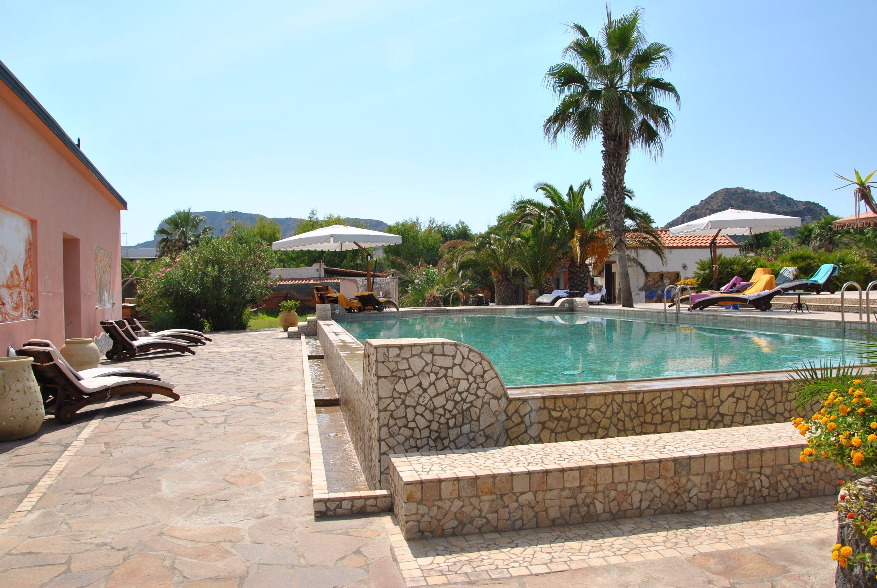 A pool area with lounge chairs, a stone wall, and palm trees.