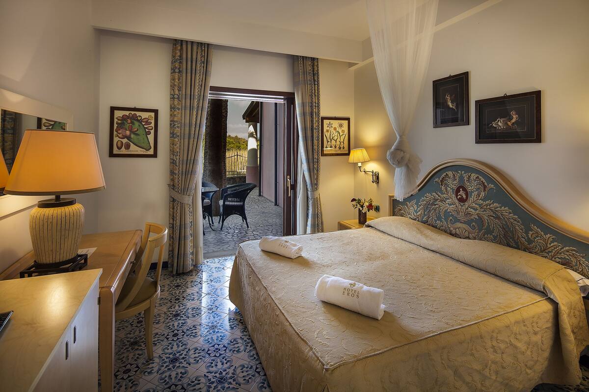 A hotel room with a bed, a desk, a chair, a lamp, and a view of the outside.