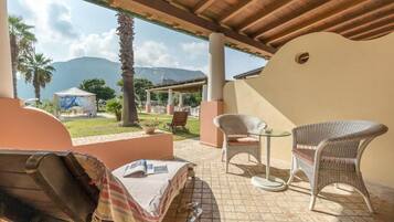 A covered outdoor seating area with wicker chairs and a table, overlooking a garden and mountains.