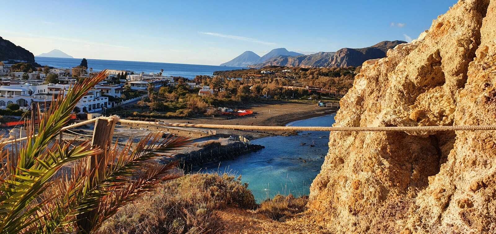 Un paese costiero con una scogliera rocciosa, un ponte sospeso e vista sul mare e montagne lontane.