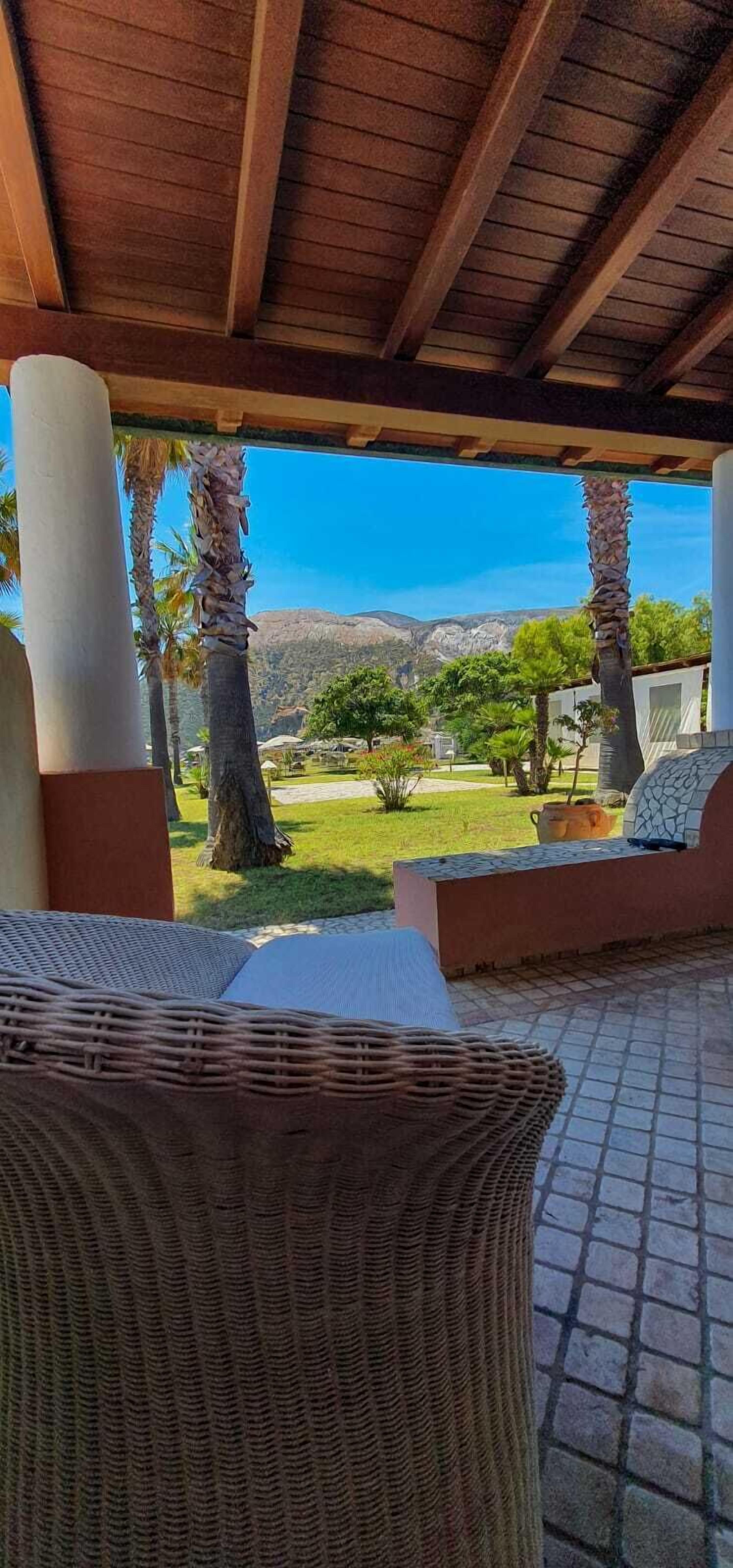 A covered outdoor seating area with wicker furniture, a view of a landscaped garden, and a mountain in the background.