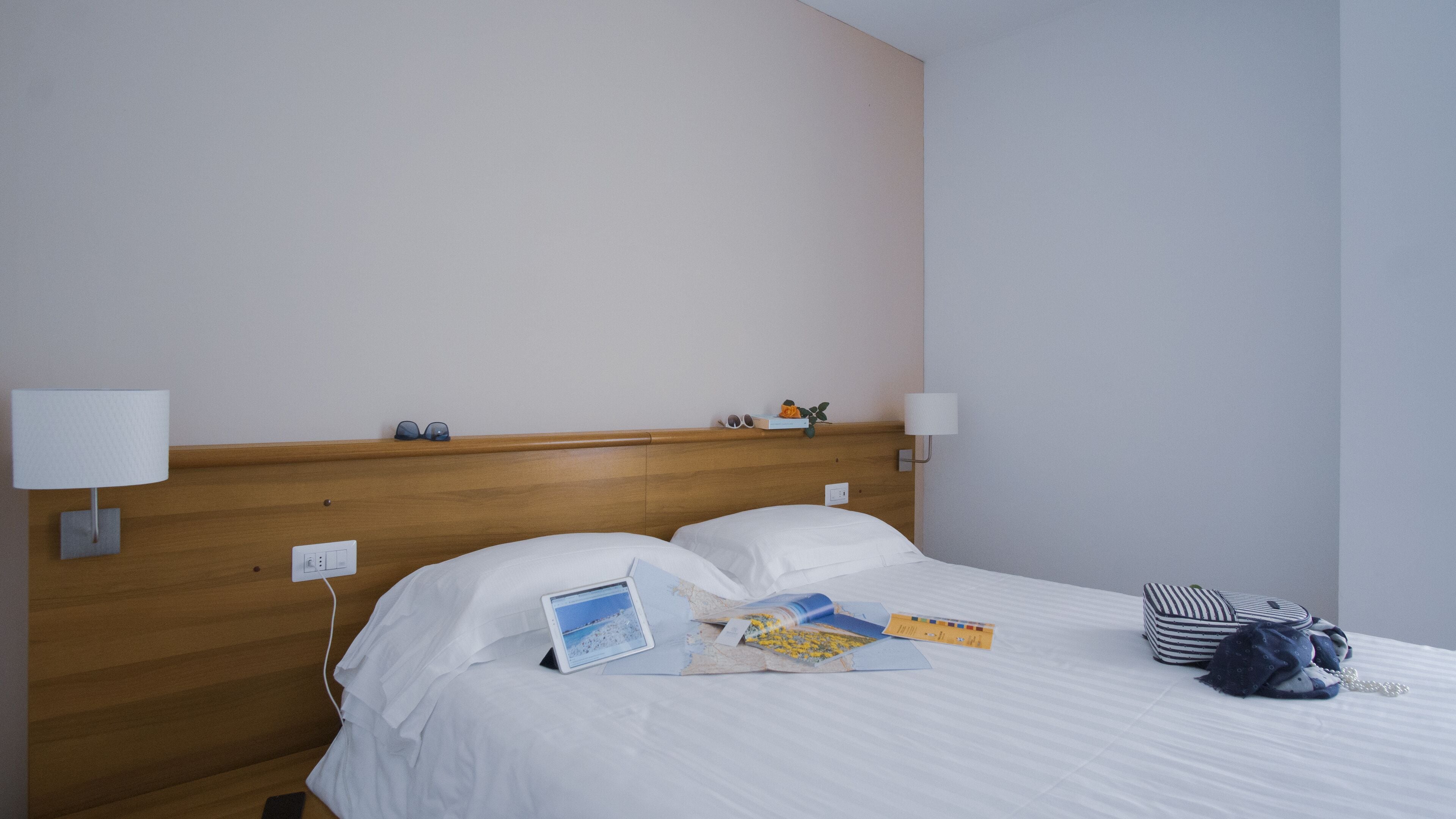 Camera d'albergo con testiera in legno, due lampade da comodino, un letto con lenzuola bianche e un piccolo tavolino con un portatile, libri e una borsa.