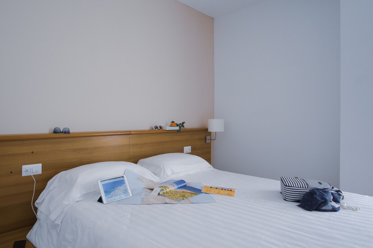 A hotel room with a wooden headboard, two bedside lamps, a bed with white linens, and a small table with a laptop, books, and a handbag.