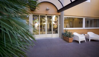 Entrada de hotel con puerta de cristal, una planta en maceta y sillas blancas afuera.