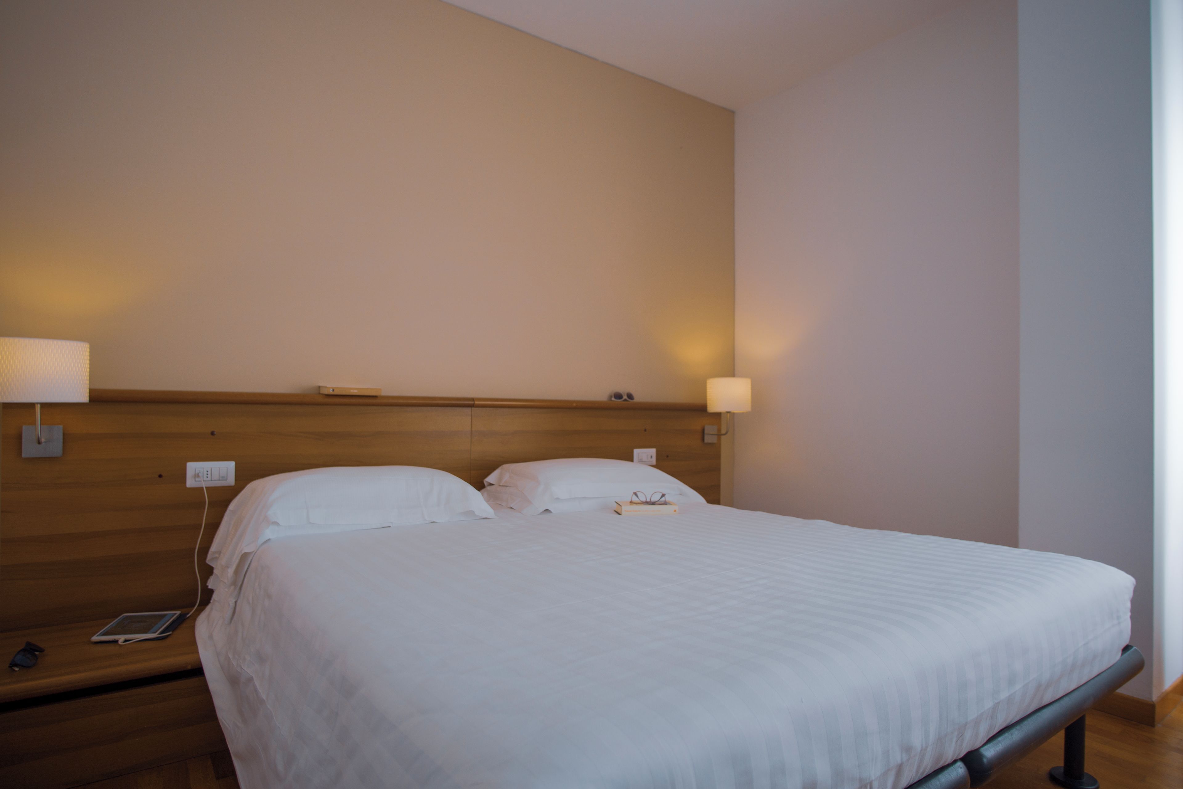 Una habitación de hotel con una cama grande, dos lámparas de noche, un cabecero de madera y una mesita de noche con un teléfono y un libro.