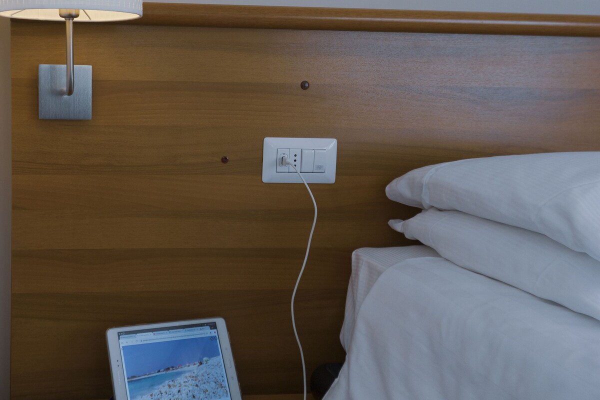 A hotel room with a wooden headboard, a bedside table with a lamp, a power outlet, a tablet, and a smartphone charger.