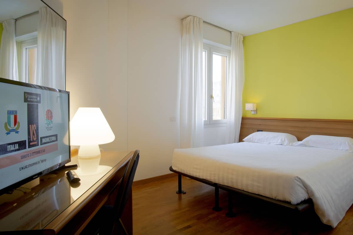 A hotel room with a bed, bedside table, lamp, TV, and a window with curtains.