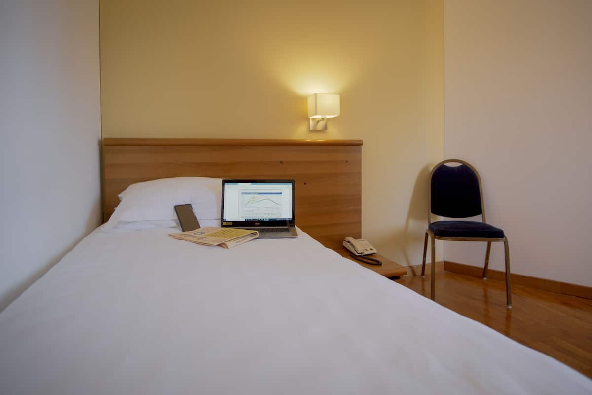 A hotel room with a bed, a laptop on the bed, a chair, and a bedside table with a phone.