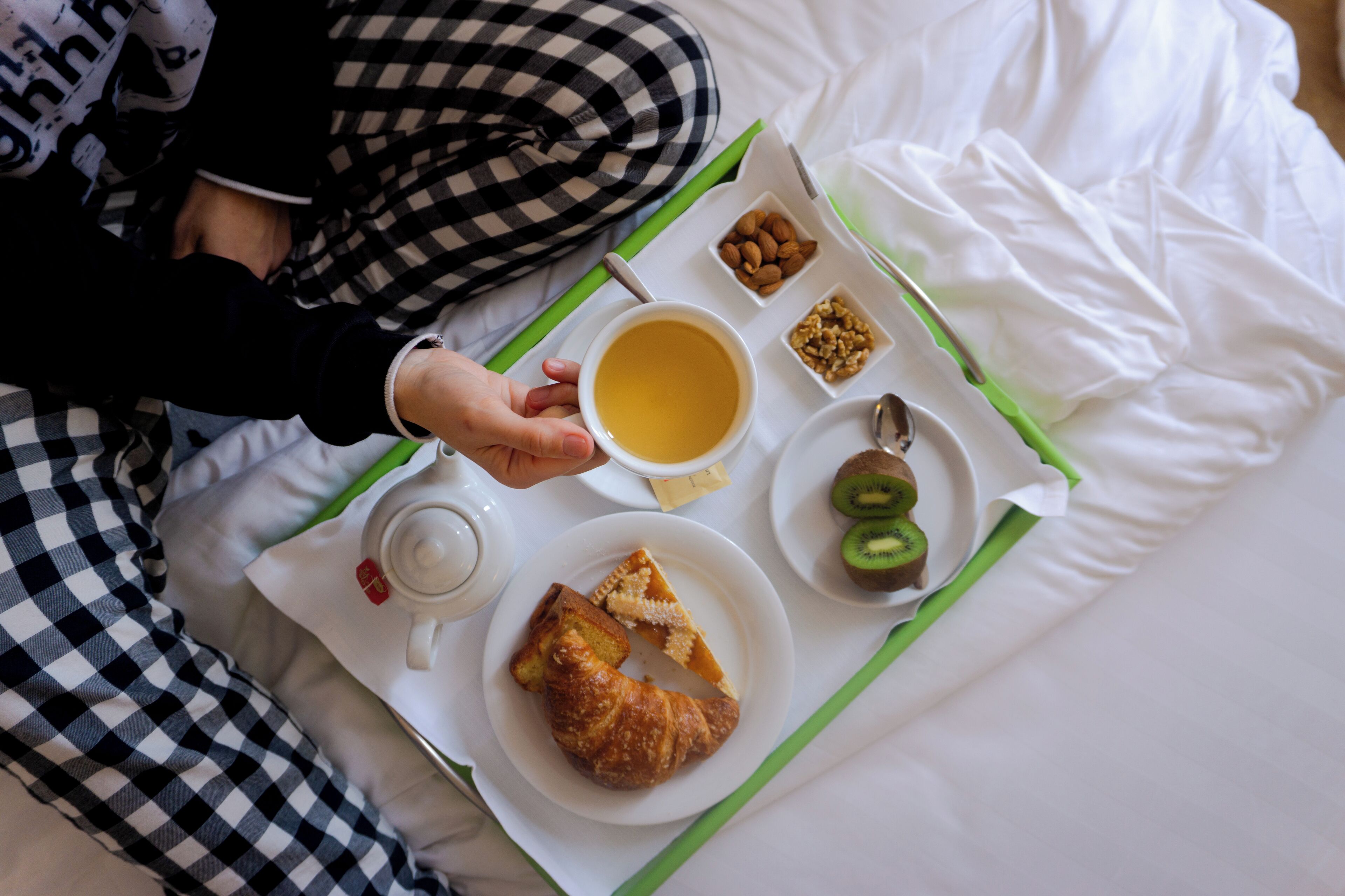 A breakfast tray with croissant, kiwi, almonds, and a cup of coffee on a bed.