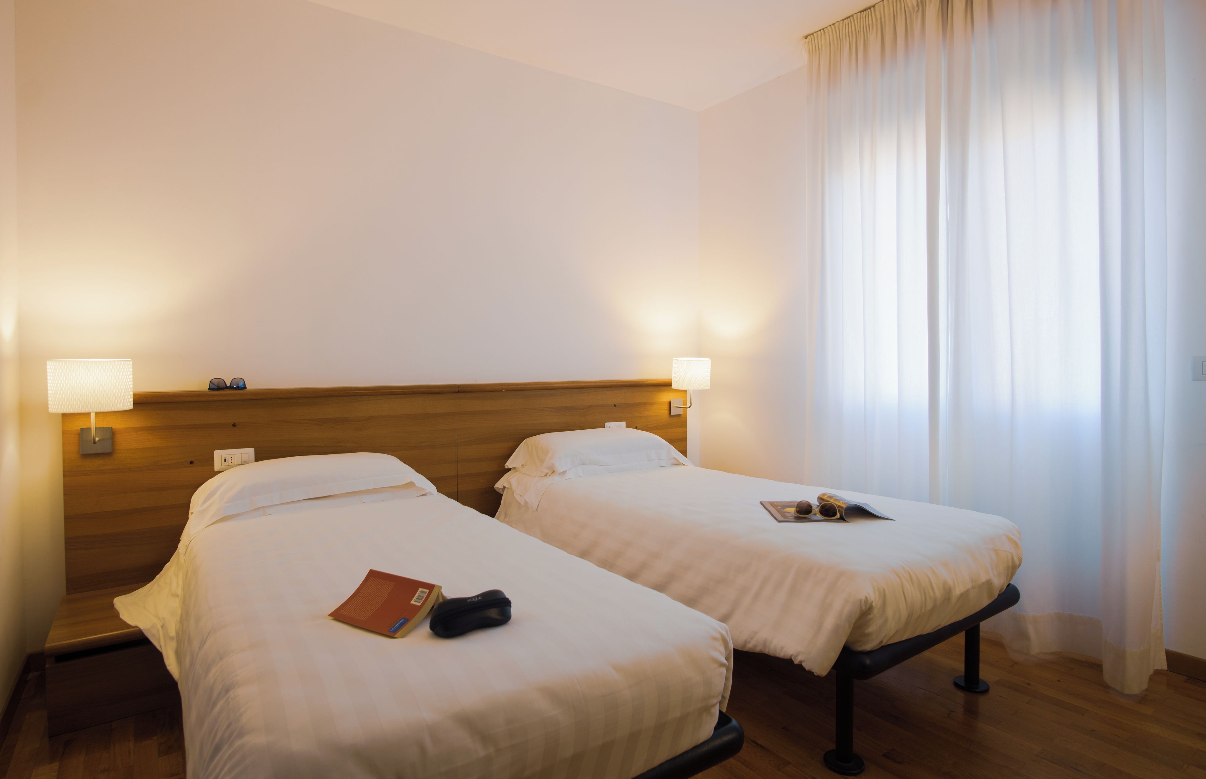 A hotel room with two single beds, a wooden headboard, bedside lamps, a wooden floor, and a window with curtains.