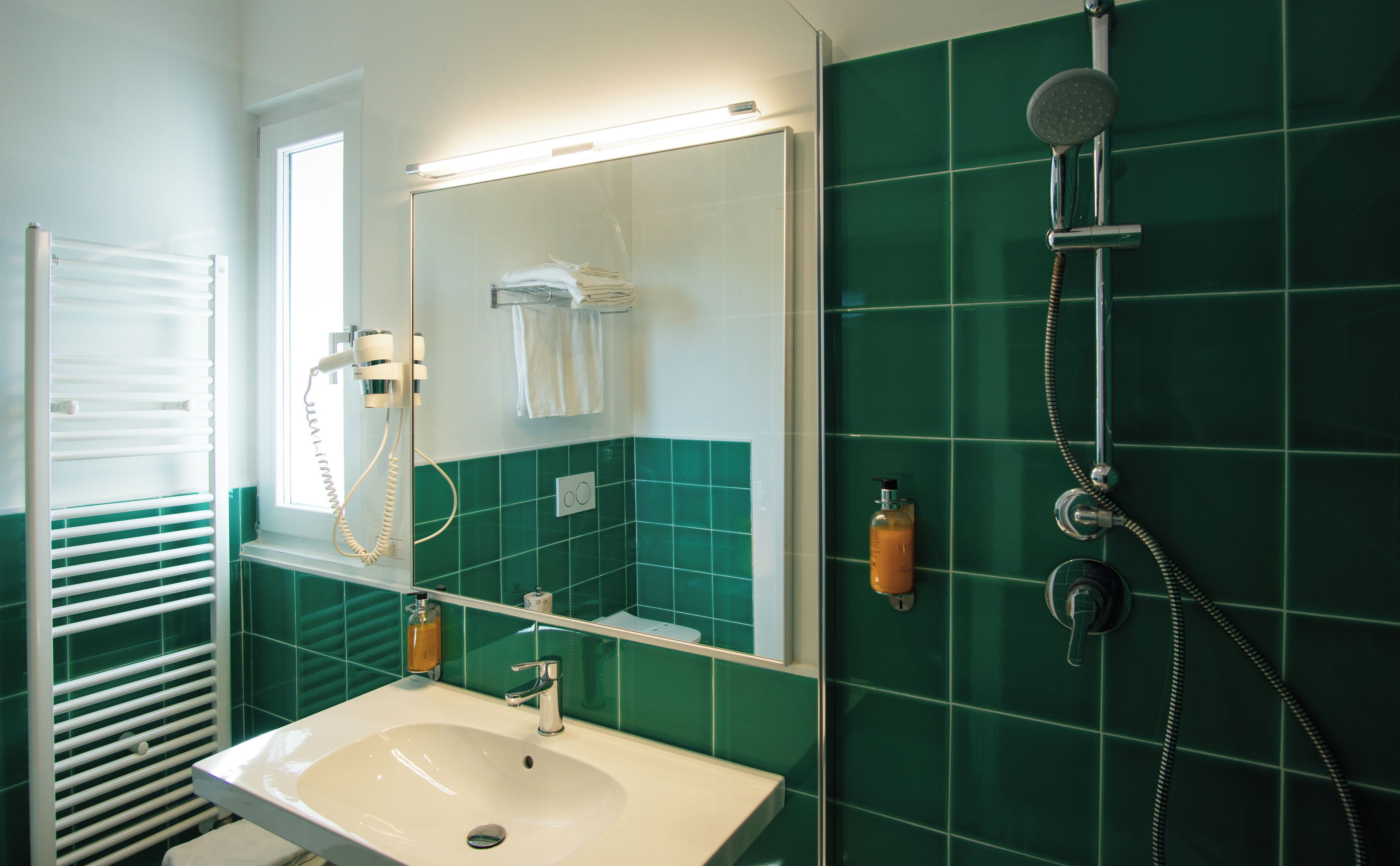 A bathroom with a white sink, a mirror, a towel rack, a shower with a handheld showerhead, and green tiles.