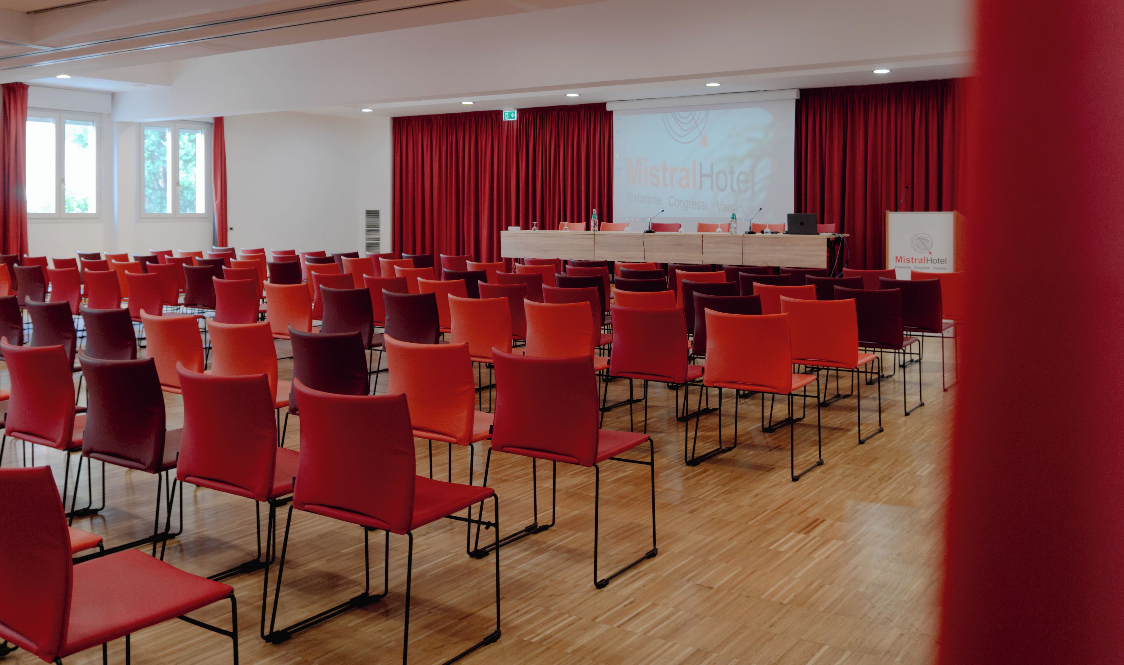 Ein Konferenzraum mit Reihen roter Stühle, einer Bühne mit einer Projektionsleinwand und einem Holztisch.
