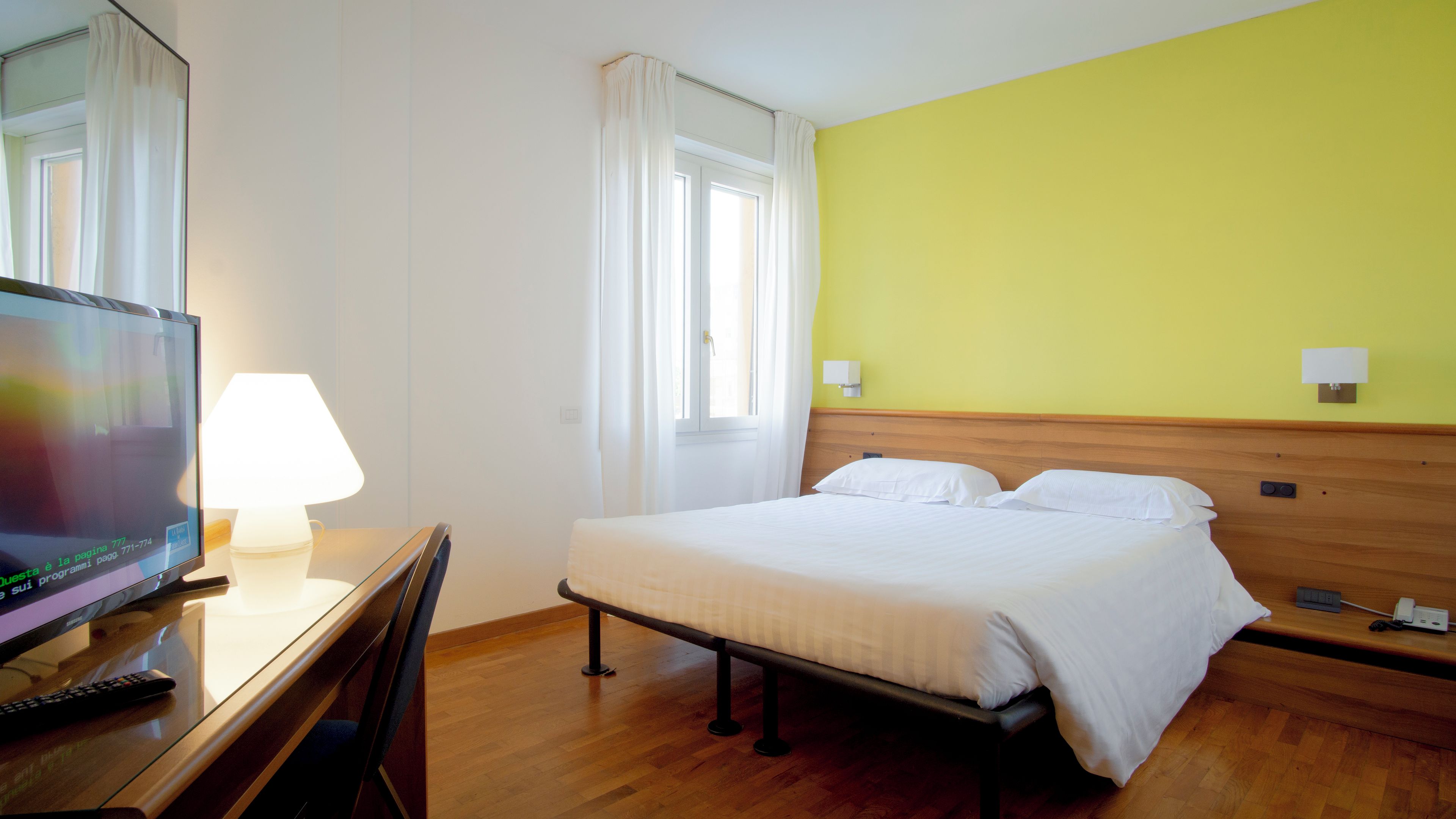 Une chambre d’hôtel avec un lit, un bureau avec une lampe, une télévision et une fenêtre avec des rideaux.