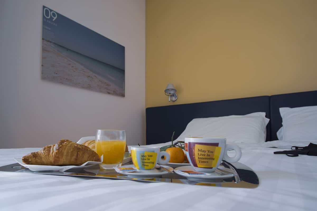 A neatly made breakfast tray with croissant, orange juice, and coffee on a hotel bed with a view of the beach and a wall-mounted lamp.