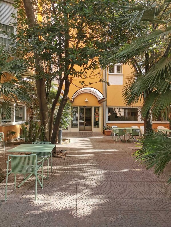 Un patio al aire libre con suelo de ladrillo, sillas y mesas de metal verde. Se ve un edificio amarillo con una entrada curva y una puerta.