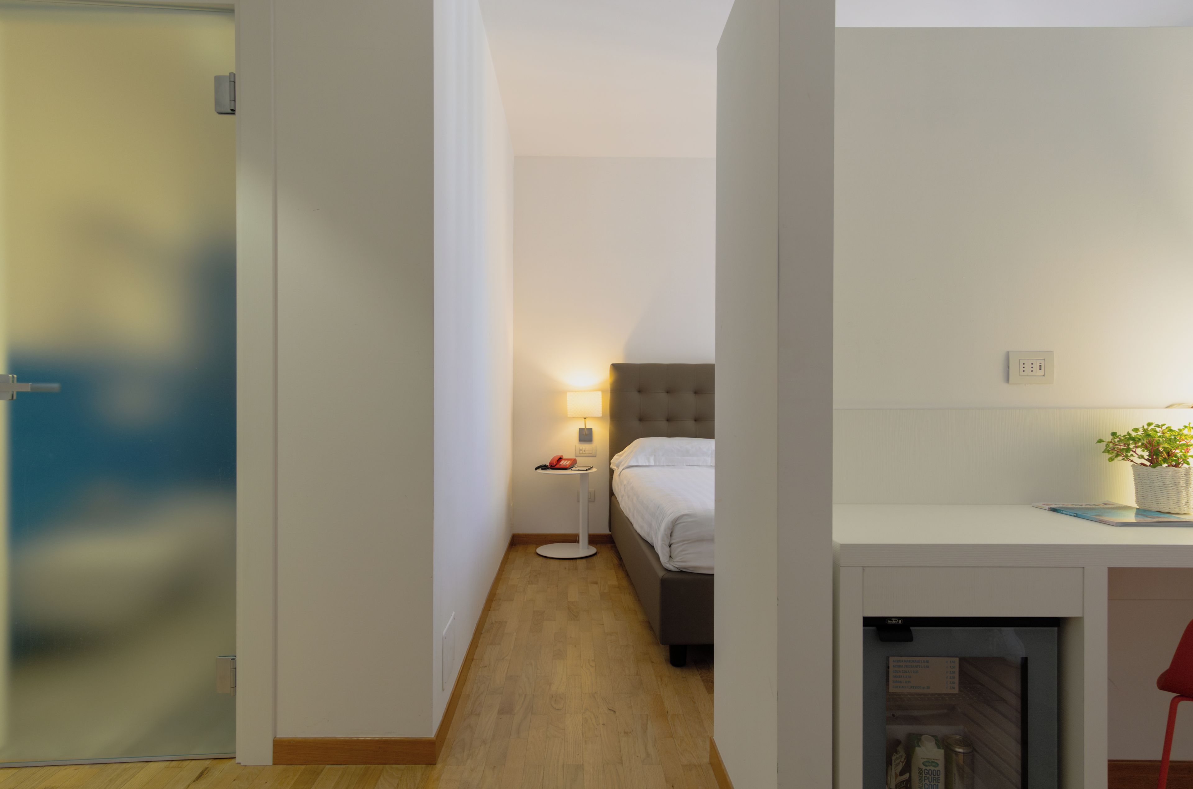 A narrow hallway leading to a bedroom with a bed and bedside table, a small table with a lamp, and a wall-mounted light fixture.