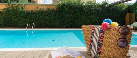 Zona junto a la piscina con una cesta de mimbre que contiene un libro, protector solar y gafas de sol.