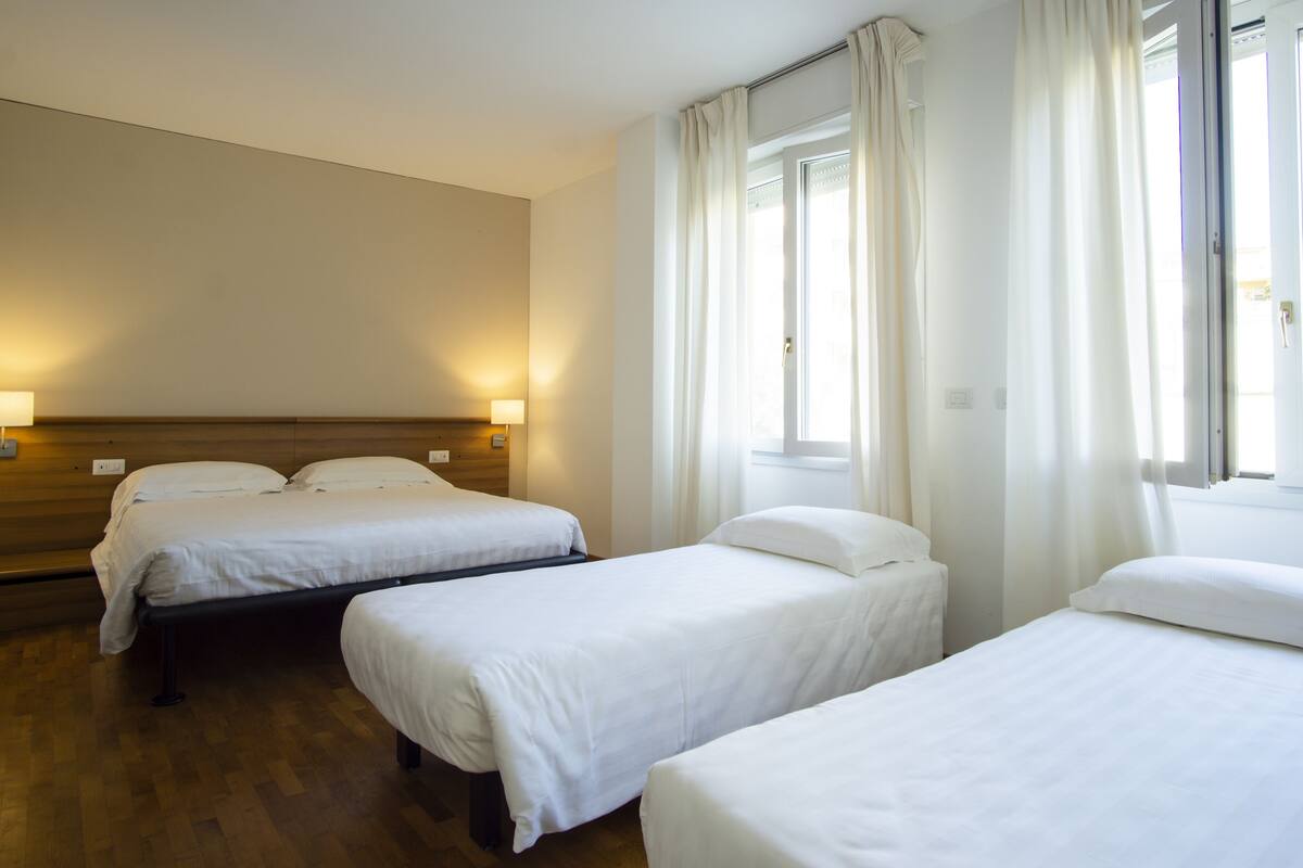 A hotel room with two single beds, a wooden headboard, bedside lamps, and a large window with curtains.