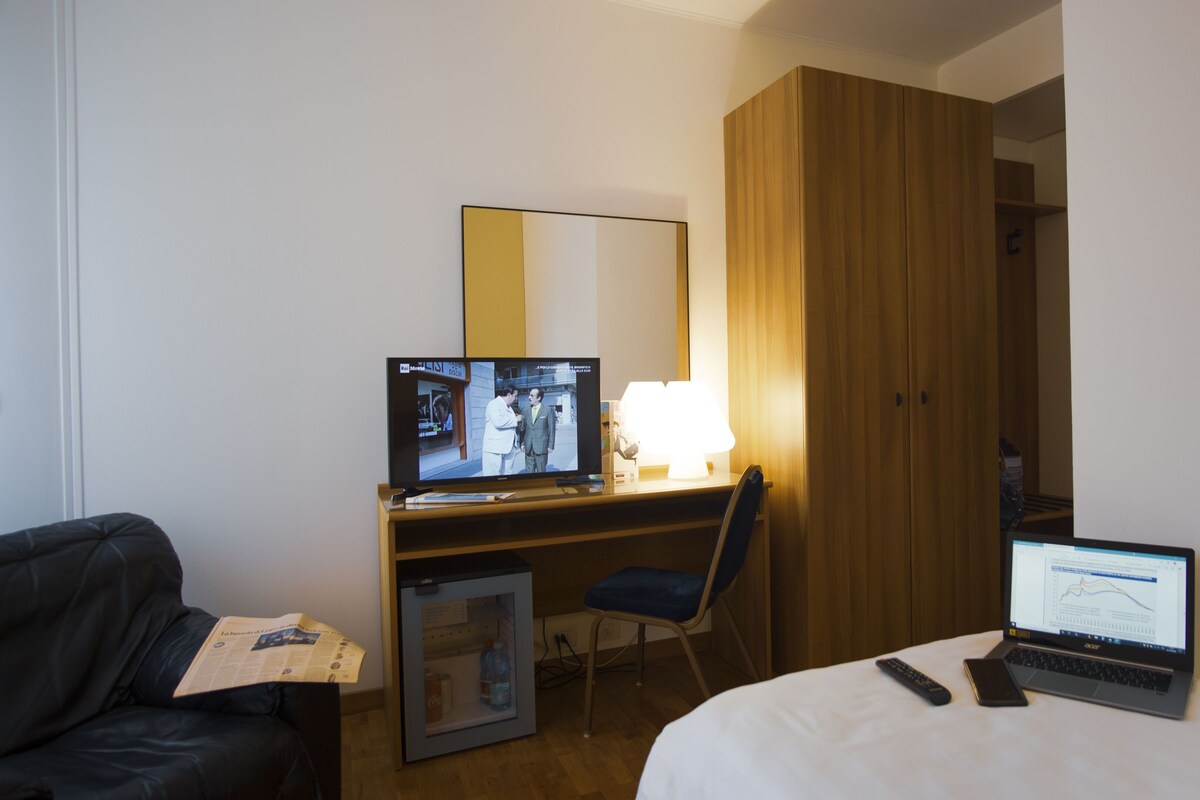 A hotel room with a bed, a desk with a computer, a chair, a TV, and a wardrobe.