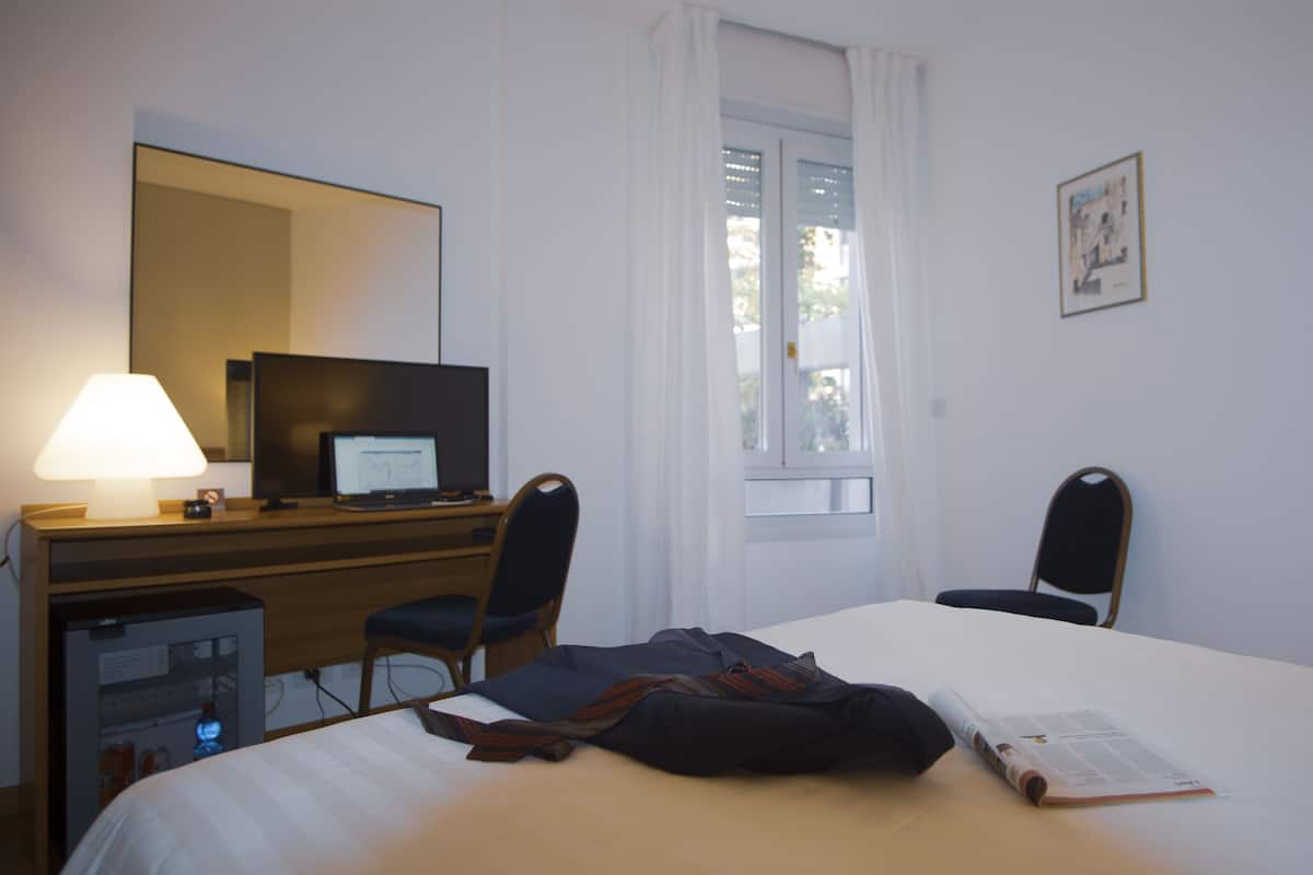 A hotel room with a bed, a desk with a laptop, a chair, a lamp, and a window with curtains.