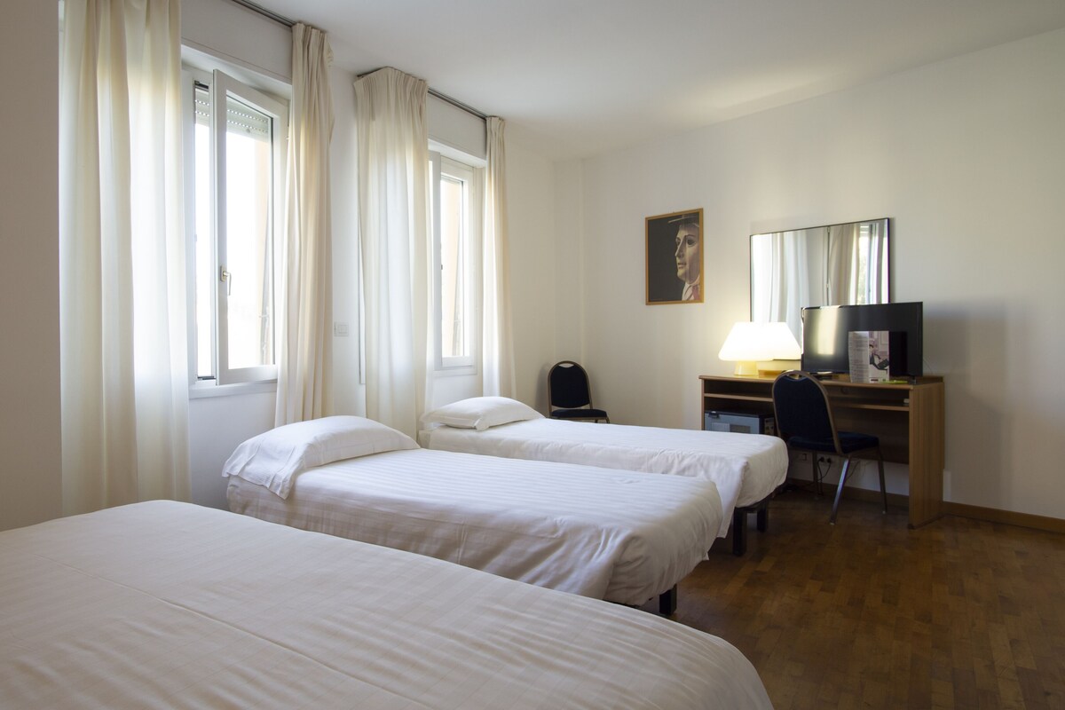 A hotel room with two single beds, a desk with a chair, a television, and a window with curtains.