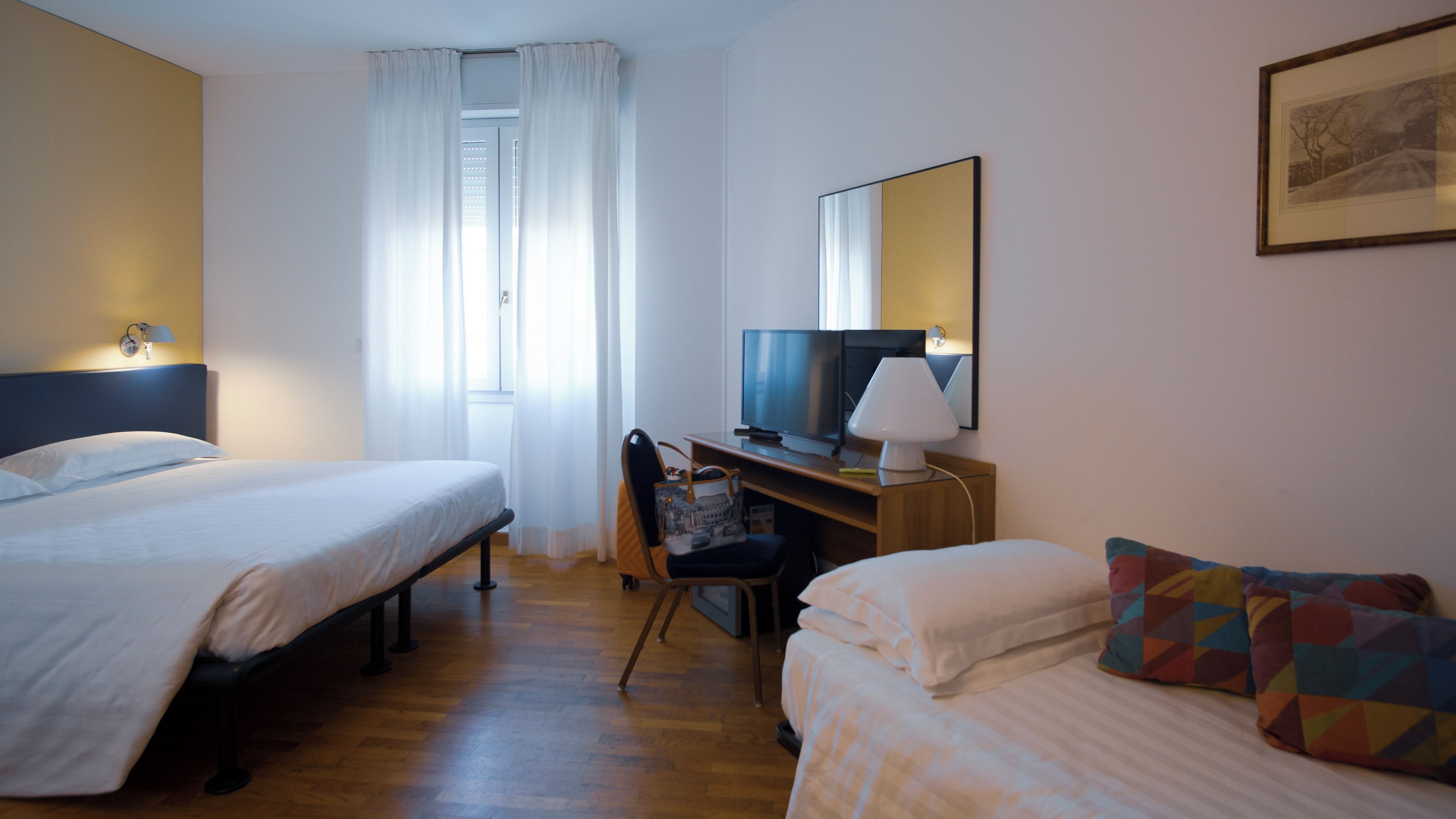 Hotellihuone, jossa on kaksi sänkyä, työpöytä, jossa on televisio, tuoli ja ikkuna, jossa on verhot.