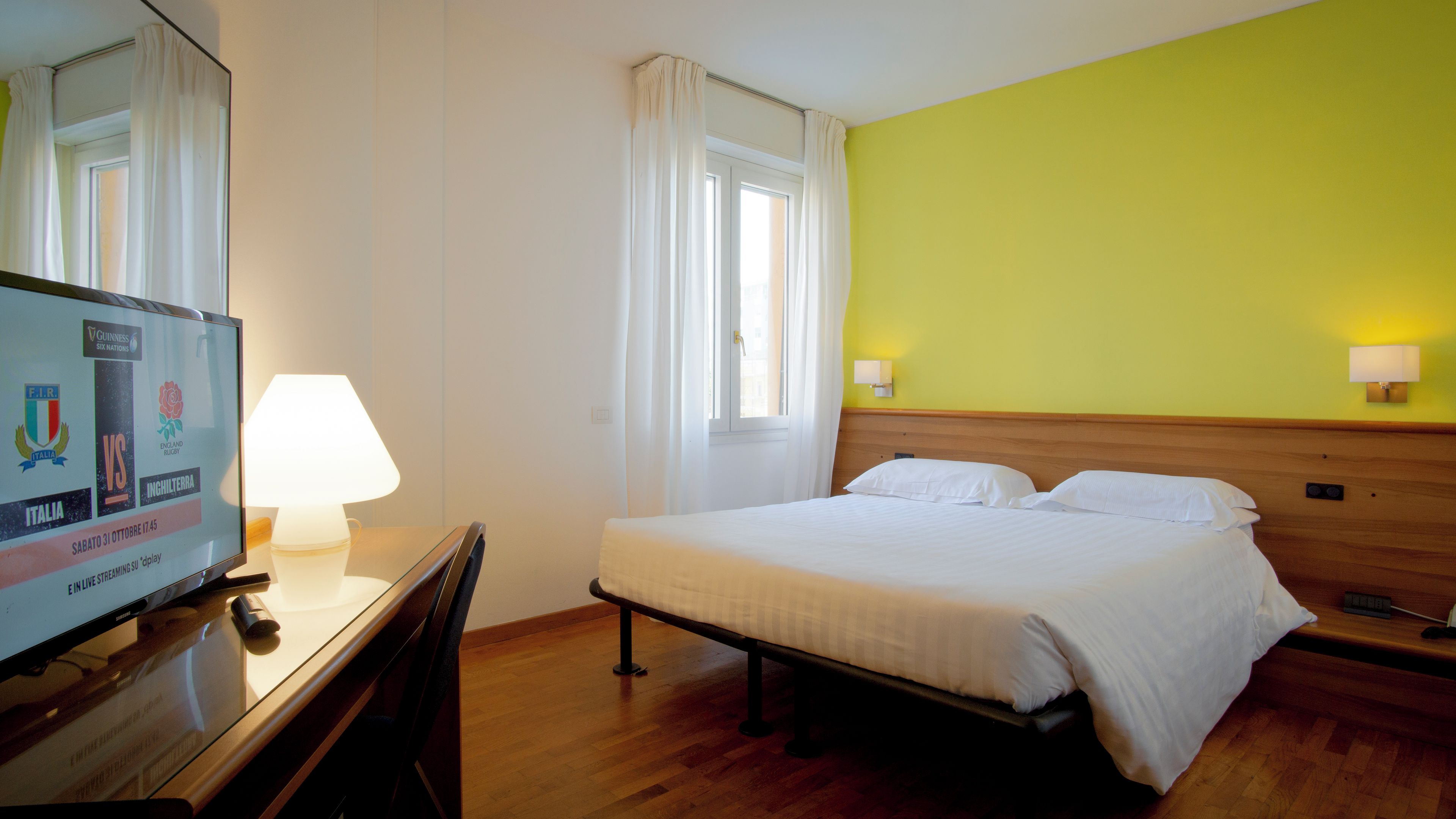 A hotel room with a bed, a desk with a lamp, a television displaying a sports event, and a window with curtains.