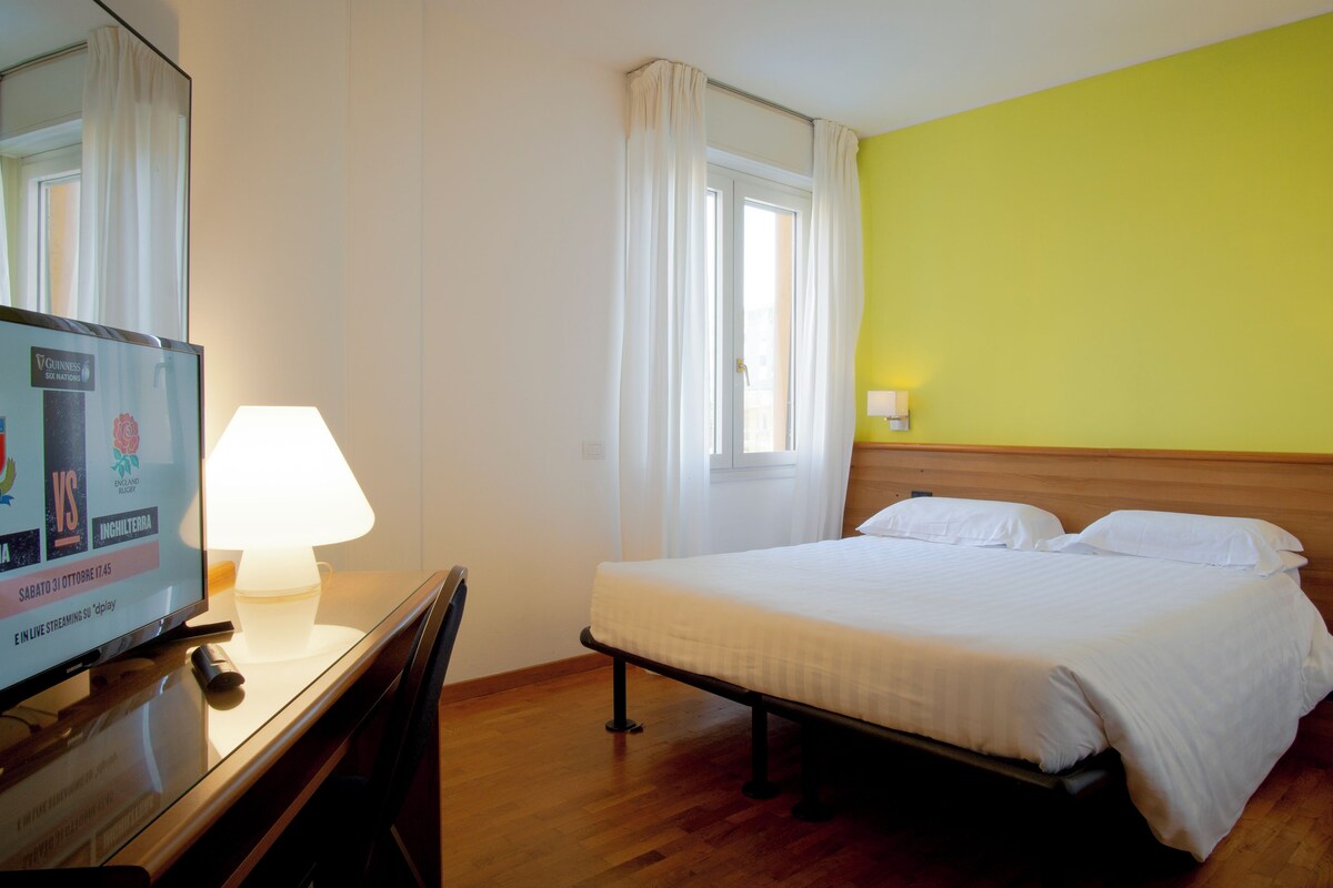 A hotel room with a bed, bedside table, lamp, TV, and a window with curtains.