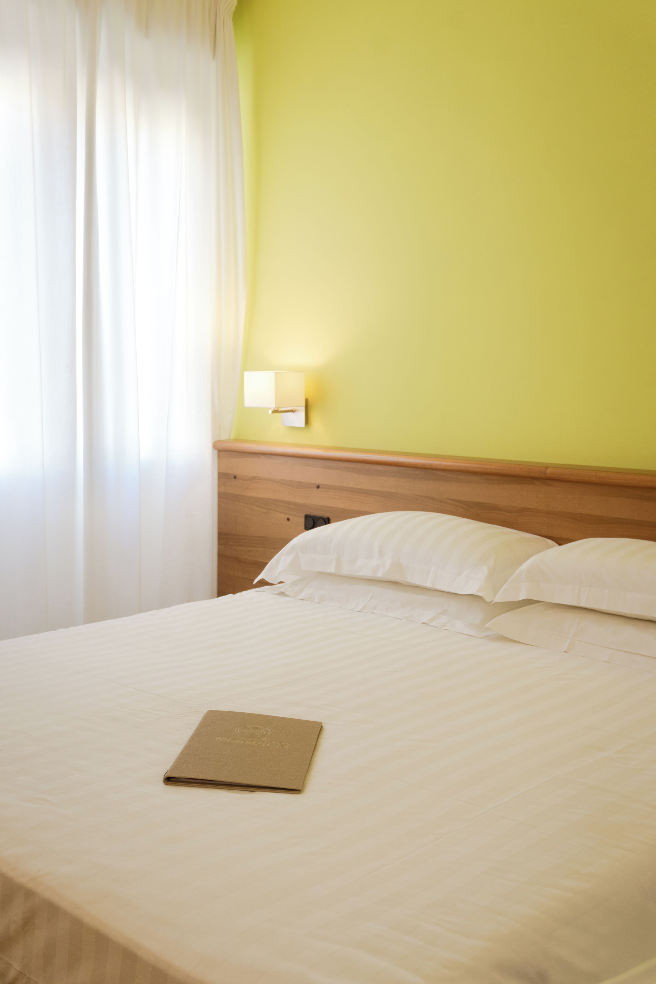 Un lit bien fait, avec un dossier marron posé dessus, un oreiller blanc et un luminaire fixé au mur.