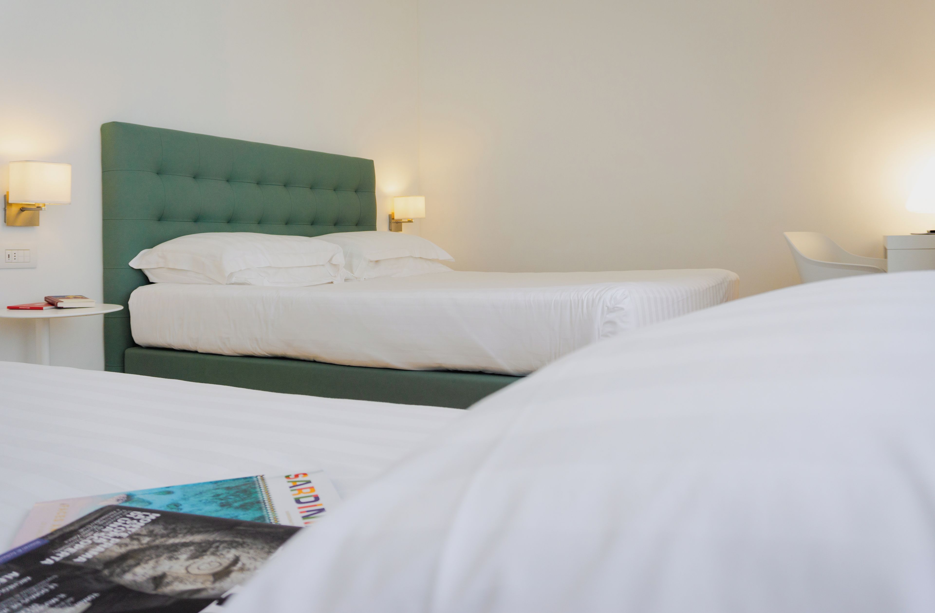A hotel room with a single bed, a green upholstered headboard, two bedside tables with lamps, and a white chair.