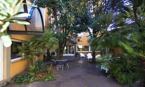 A courtyard with a table and chairs, surrounded by palm trees and a building with a glass entrance. - Hotel Mistral 2 (Oristano)