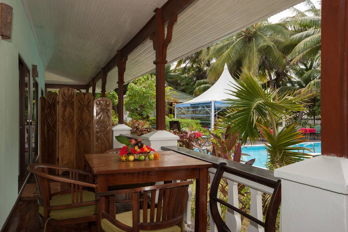 Uma mesa de madeira com cadeiras e uma fruteira, uma cerca branca e uma piscina com palmeiras ao fundo.