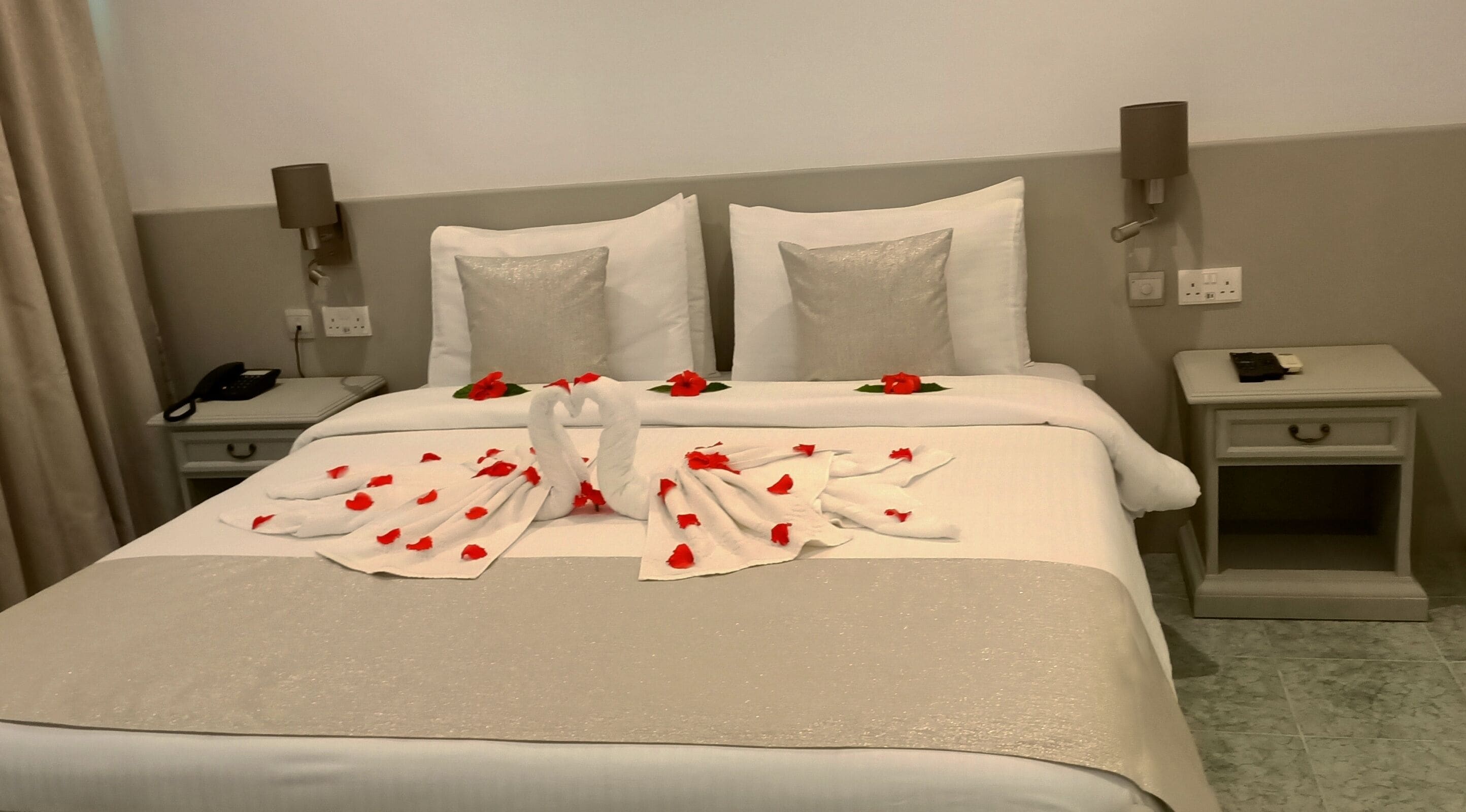 A neatly made bed with white linens, a swan-shaped towel arrangement, and red rose petals. There are two bedside tables, one with a phone and the other with a lamp.