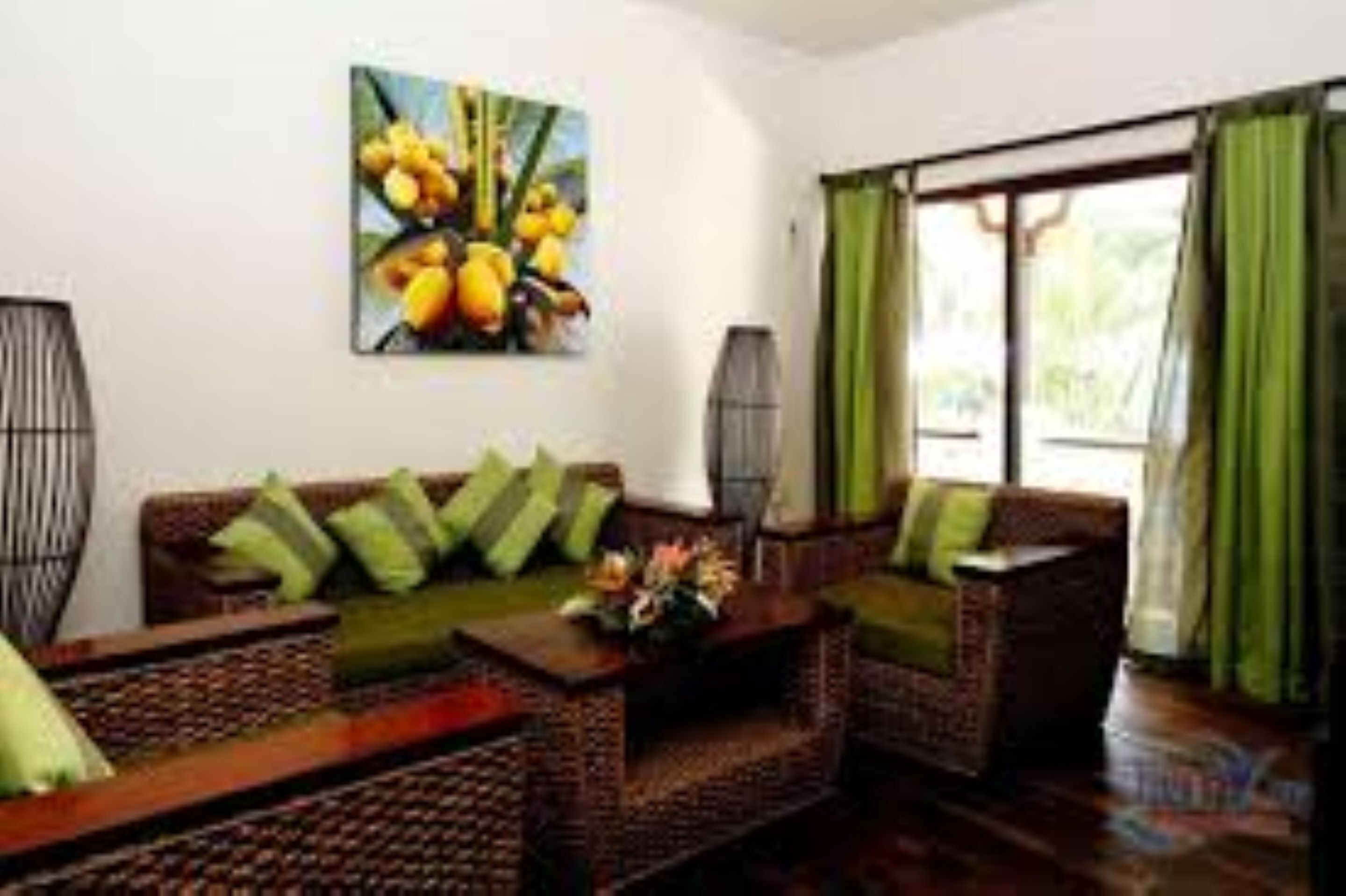 Un salón con muebles de mimbre, un cuadro de frutas y cortinas verdes.