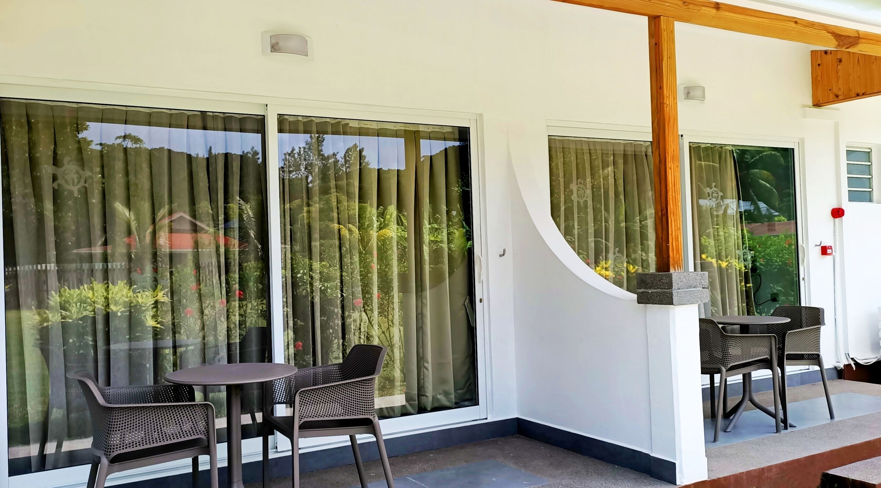 Un espace extérieur abrité, doté de deux tables et de chaises, de grandes fenêtres avec des rideaux et d’une poutre en bois au plafond.