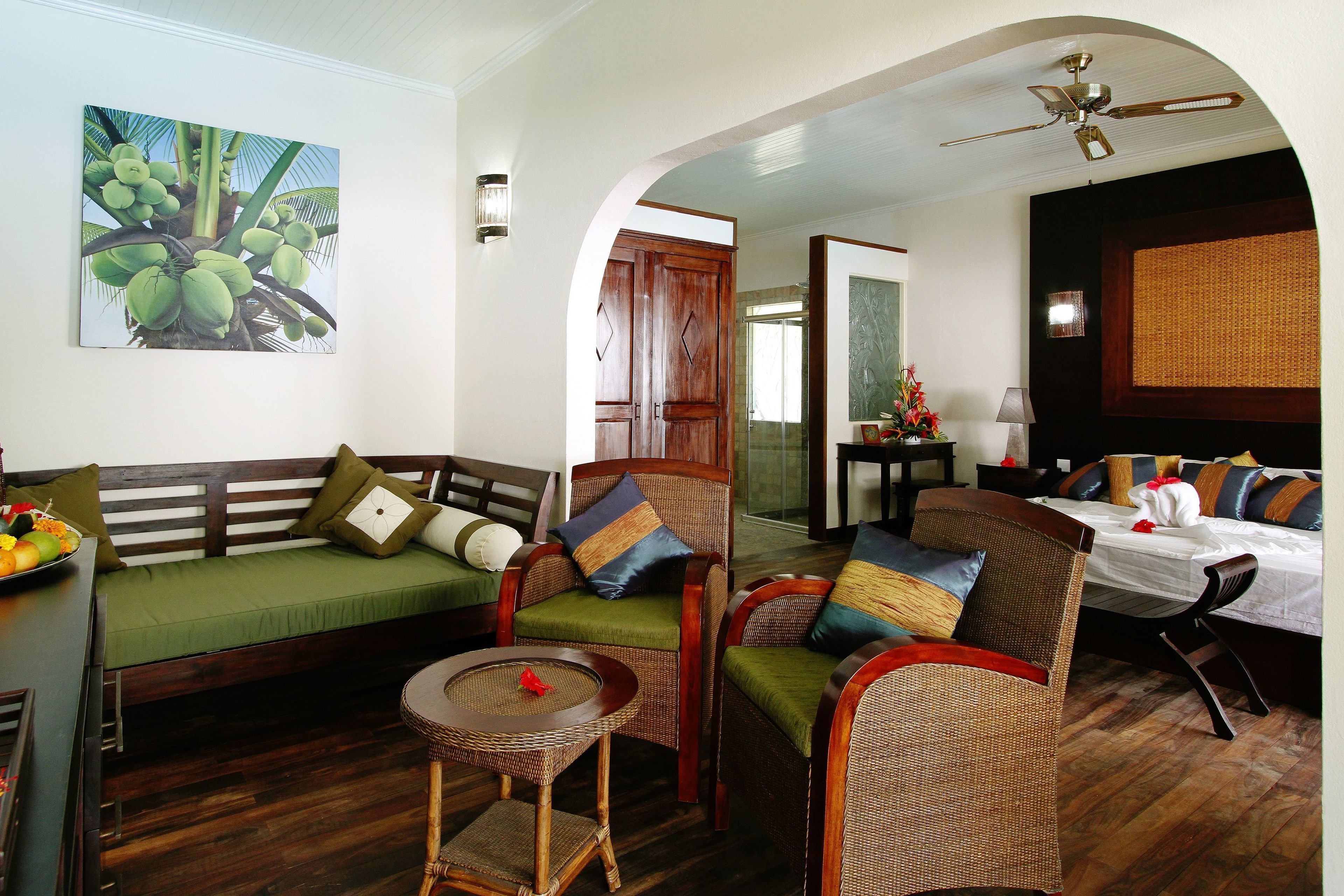 A room with a sofa, chairs, a small table, a bed, and a ceiling fan.