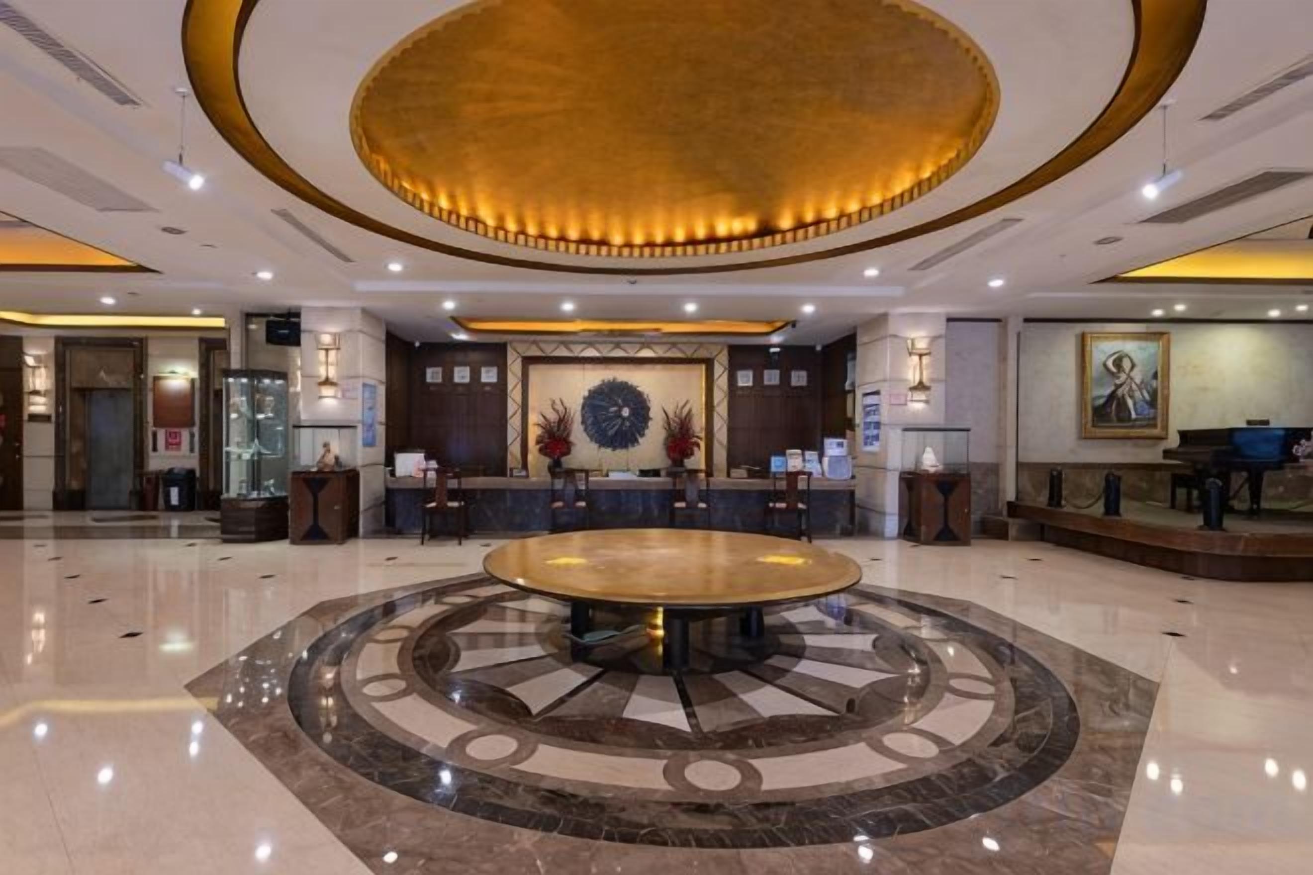 A hotel lobby with a central round table, reception desk, and a decorative wall.