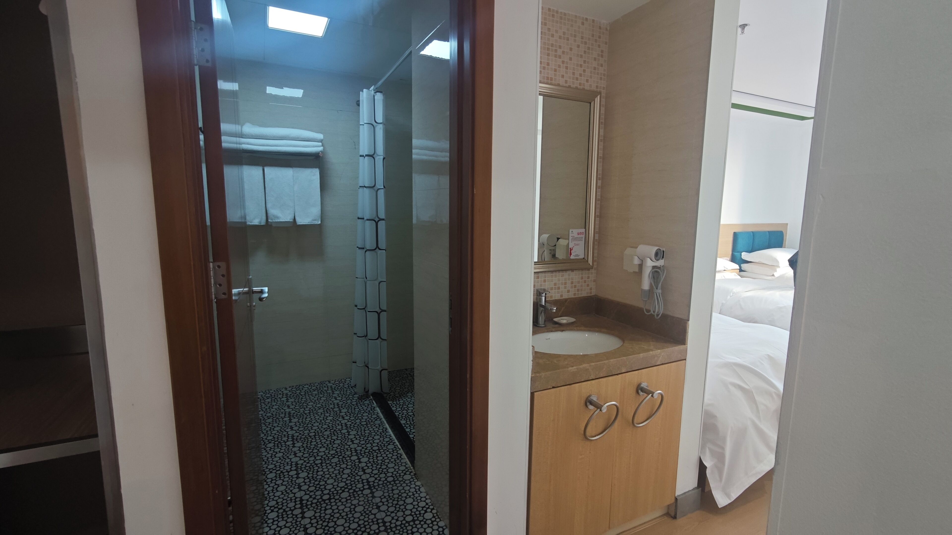 A hotel room with a glass door leading to a bathroom, a bed with white bedding, a wooden cabinet with two drawers, a round sink, and a mirror above the sink.