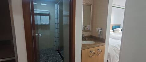 A hotel room with a glass door leading to a bathroom, a bed with white bedding, a wooden cabinet with two drawers, a round sink, and a mirror above the sink.