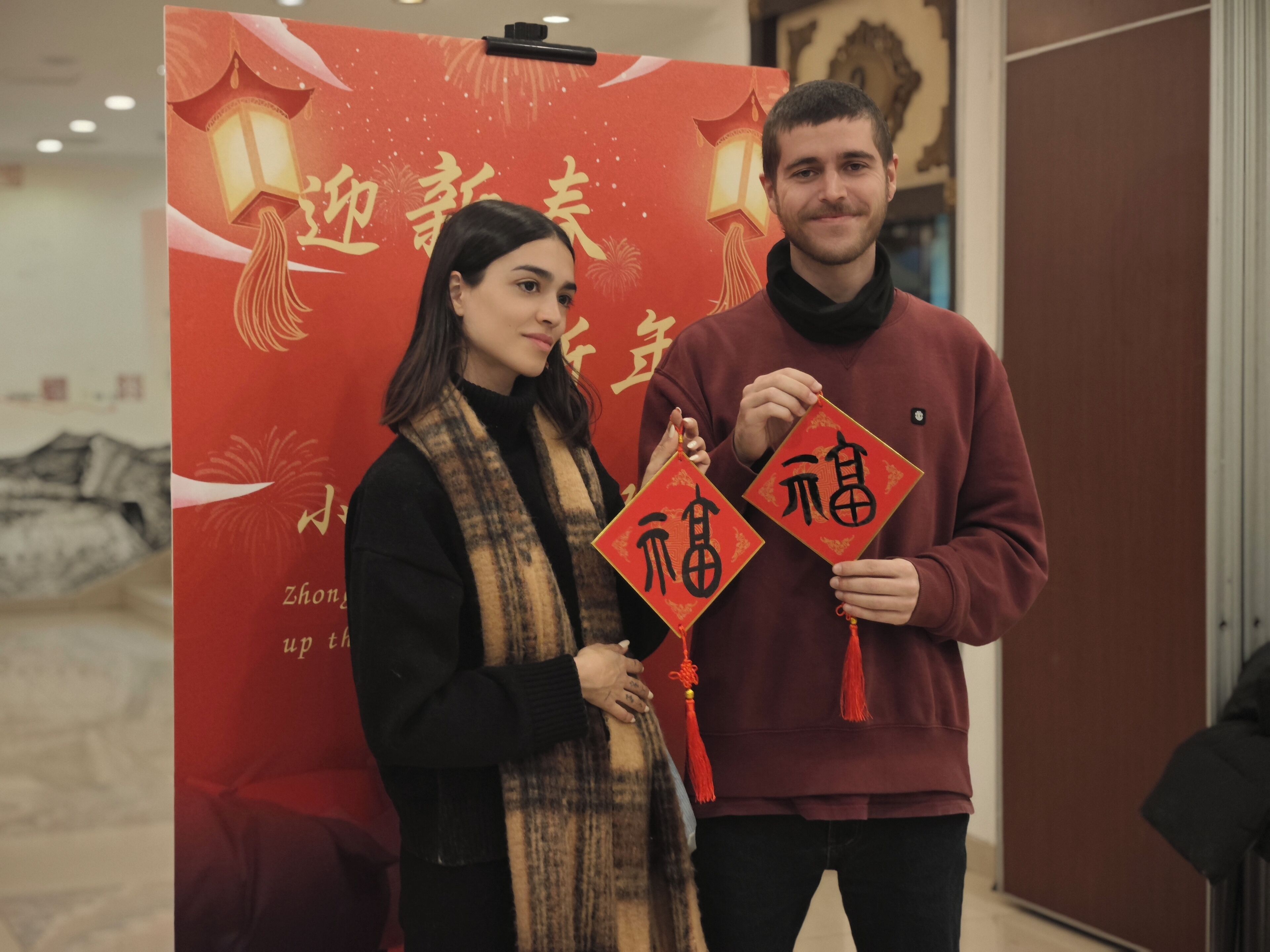 Ein Mann und eine Frau halten rote Laternen mit chinesischen Schriftzeichen vor einem roten Hintergrund mit Feuerwerk und chinesischem Text.