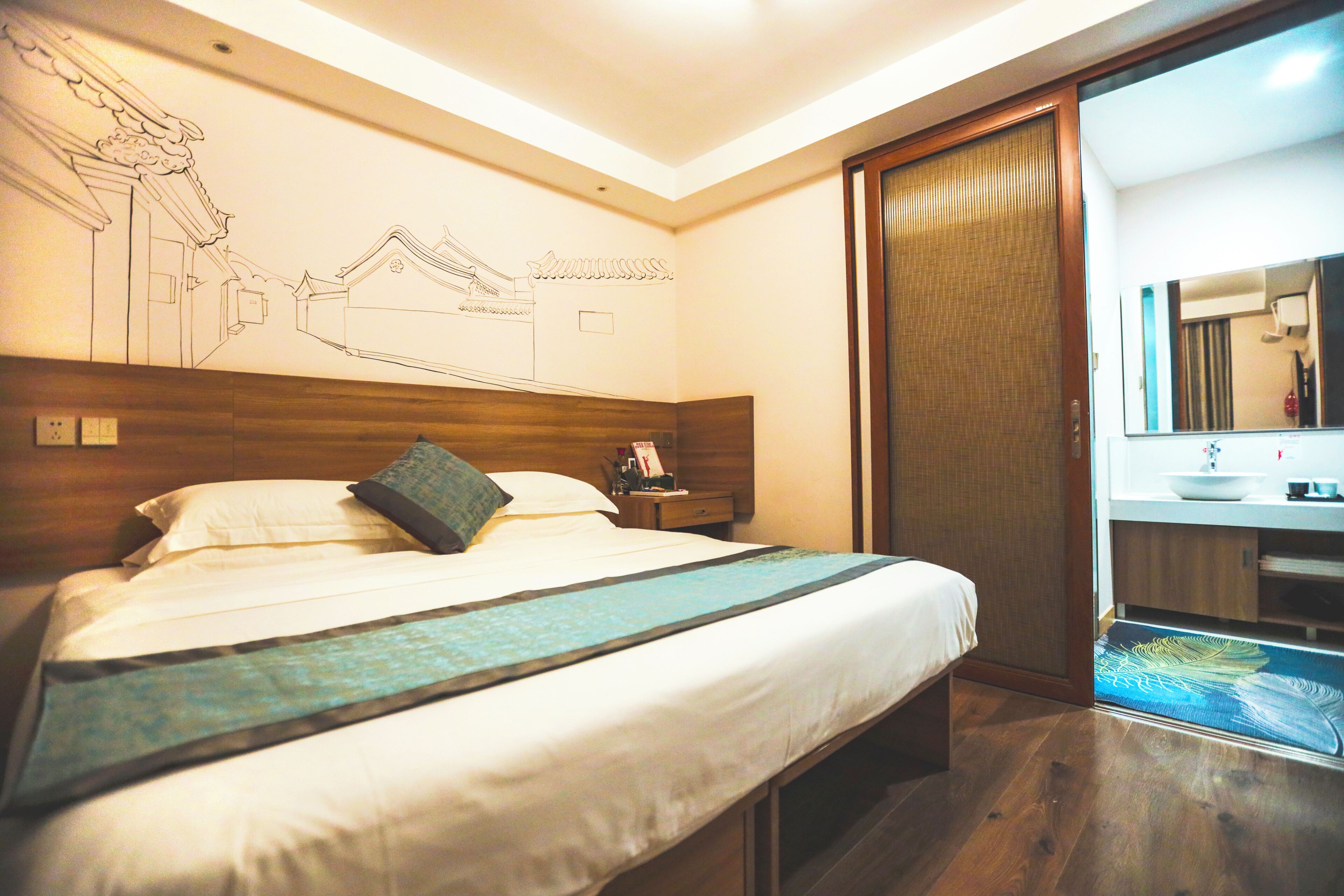 A hotel room with a bed, wooden headboard, a nightstand, a mirror, and a bathroom visible through an open door.