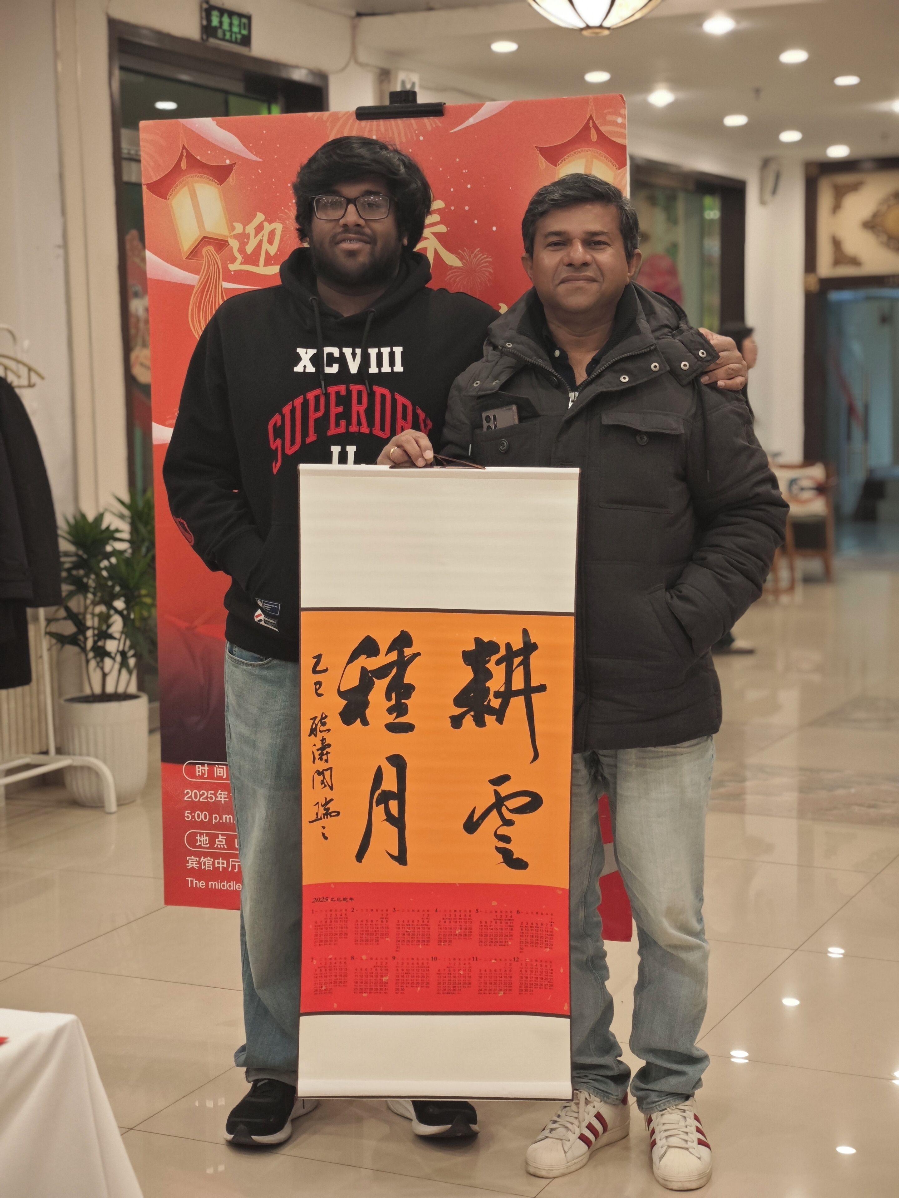 Two people standing in front of a red and white poster with Chinese characters.